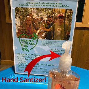 poster for "Hearts of Glass" event on a table with hand sanitizer; red arrow points with text "hand sanitizer!"
