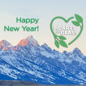 Square graphic. Photo of the Teton Mountain Range at sunset. The snow-covered peaks have pink highlights. Green text "Happy New Year!" Next to the text is the logo for the film "Hearts of Glass."